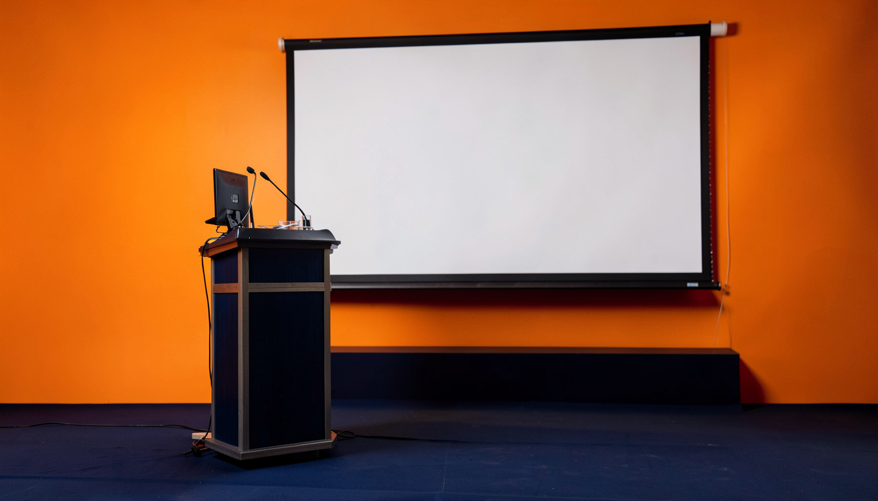 A clean academic conference setting with deep navy and soft gold tones, featuring a podium and large projection screen
